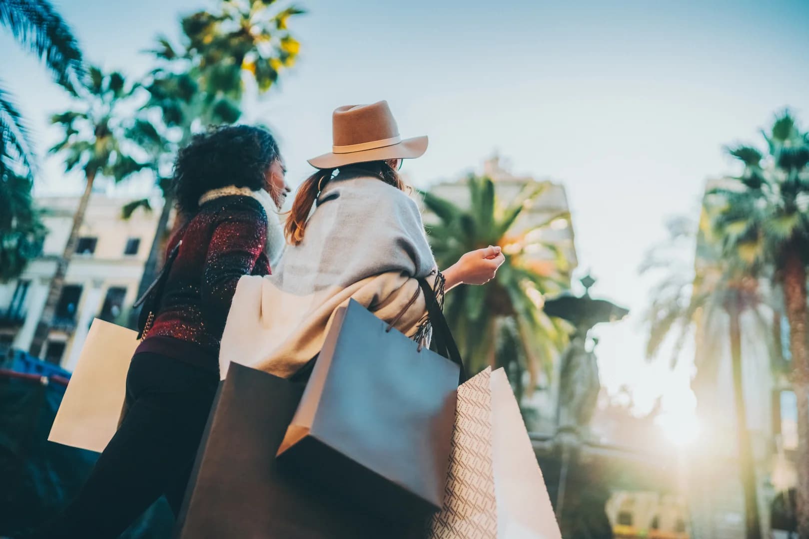 Two Women Shopping at an Outdoor Plaza with Palm Trees and Shopping Bags